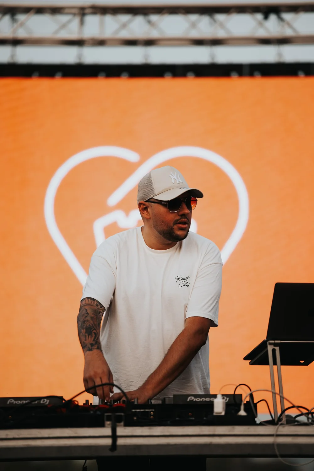 A DJ at the decks in front of the stage screen, which shows the mark in white on orange